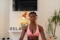 Femme afro-américaine détendue assise dans Lotus pose et écoute la médiation dans les écouteurs tout en faisant du yoga à la maison — Photo de stock