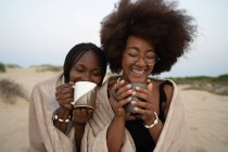 Счастливые молодые чернокожие подруги с кружками горячих напитков сидят рядом, завернутые в теплое одеяло, и веселятся летним вечером на песчаном пляже — стоковое фото