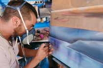 Vista laterale dell'artista maschio in respiratore utilizzando pistola a spruzzo per dipingere quadro su tela durante il lavoro in laboratorio creativo — Foto stock