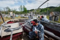 Unrecognizable men with guns and in protective helmets playing paintball among abandoned boat and cars — Stock Photo