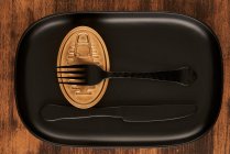 Vista superior de tenedor negro y cuchillo colocado cerca de la comida enlatada sellada en bandeja negra rectangular - foto de stock