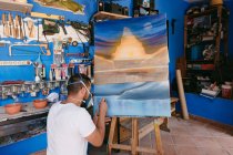 Hombre en respirador rociando pintura sobre lienzo con paisaje abstracto mientras trabaja en un estudio creativo profesional - foto de stock