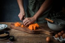 Unrecognizable male cook in apron chopping raw pumpkin on wooden cutting board near flour and bread crumbs with seeds and eggs while preparing pie — Stock Photo