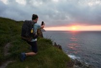 Обратный вид на неузнаваемого мужчину-путешественника, фотографирующего закат над морем на смартфоне летом — стоковое фото
