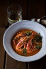 From above of delicious spicy stew with chickpeas and big shrimps decorated with cilantro leaves served in white soup plate on wooden table with glass of drink and spoon — Stock Photo