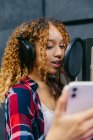 Alegre cantante afroamericana femenina en auriculares con smartphone interpretando canción contra escudo de sonido en estudio de grabación - foto de stock