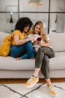 Corpo pieno allegra coppia lesbica in abiti casual navigazione cellulare e seduti insieme sul comodo divano in soggiorno moderno — Foto stock
