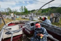 Homens irreconhecíveis com armas e em capacetes de proteção jogando paintball entre barco abandonado e carros — Fotografia de Stock