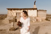 Woman in white dress and with flowers standing against shabby aged cabin on wedding day in Bardenas Reales Natural Park in Navarra, Spain — Stock Photo