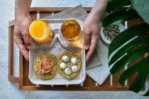 Top view of crop unrecognizable person eating appetizing healthy avocado toasts with salmon and burrata cheese served on tray with glasses of fresh juices in sunlight — Stock Photo