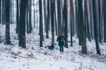 Леди в лыжной куртке гуляет с домашней собакой между деревьями в зимнем лесу — стоковое фото