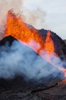 Всплески горячей апельсиновой лавы, извергающейся с вулканической вершины горы, окруженной дымом в Исландии — стоковое фото