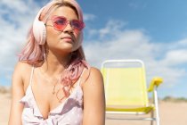 Mujer serena con cabello rosa escuchando música en auriculares mientras se enfría en la orilla del mar en un día soleado en verano - foto de stock