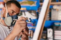 Vue latérale de l'artiste masculin dans un respirateur à l'aide d'un pistolet pulvérisateur pour peindre sur toile pendant le travail dans un atelier créatif — Photo de stock