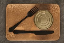 Верхний вид черной вилки и ножа помещен рядом с герметичной консервной пищей на прямоугольный медный поднос — стоковое фото