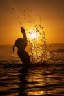 Side view silhouette of anonymous little girl standing in sea water and making splashes with hands against sunset light in summertime — Stock Photo