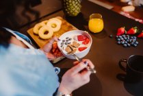 Высокий угол урожая неузнаваемая женщина сидит и ест вкусные хлопья с йогуртом и ягодами утром — стоковое фото