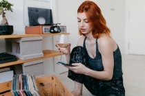 Side view of young female with glass of alcoholic drink listening to song from vintage record player and browsing phone — Stock Photo