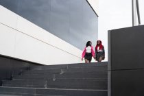 Nível do solo corpo cheio de mulheres afro-americanas em desgaste elegante fazendo passarela caminhar juntos perto do edifício moderno — Fotografia de Stock