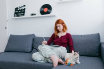 Молодая босиком женщина с рыжими волосами просматривает интернет на планшете, сидя с котом на диване дома — стоковое фото