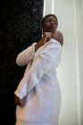 De baixo afro-americano fêmea em branco vestido da moda de pé perto da parede no quarto e olhando para longe — Fotografia de Stock