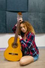 Young ethnic female guitarist with curly hair leaning on acoustic guitar while looking at camera — Stock Photo