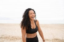 Atleta asiática positiva con el pelo rizado riéndose en la orilla del mar arenosa en verano - foto de stock
