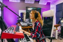 Vista lateral da cantora afro-americana sorridente no fone de ouvido tocando sintetizador enquanto executa música no microfone no estúdio de gravação — Fotografia de Stock