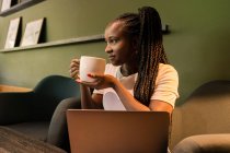 Delighted African American female sitting in cafe with cup of coffee and enjoying weekend while looking away — Stock Photo