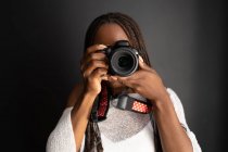 Afroamerikanische Fotografin mit Zöpfen blickt durch Fotos, die auf professioneller Kamera aufgenommen wurden, während sie auf schwarzem Hintergrund steht — Stockfoto