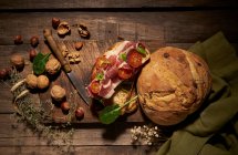 Вид на аппетитный домашний сэндвич из свежеиспеченного ремесленного хлеба с нарезанной ветчиной и помидорами на деревенском деревянном столе с орехами и травами — стоковое фото