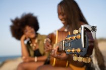 Fröhliche junge afroamerikanische Freundinnen spielen Gitarre, während sie zusammen am Sandstrand sitzen und die Sommerferien genießen — Stockfoto