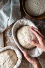 Mãos da mulher De cima segurando pão sourdough no suporte de vime com pano na mesa de madeira com farinha espalhada — Fotografia de Stock