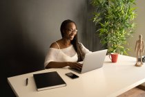 Allegra freelance afroamericana seduta a tavola sul posto di lavoro moderno e navigando in netbook mentre lavora su un progetto remoto da casa — Foto stock