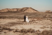 Vue arrière de mariés méconnaissables en vêtements élégants marchant sur une plaine aride vers les montagnes lors de la célébration de mariage dans le parc naturel de Bardenas Reales en Navarre, Espagne — Photo de stock