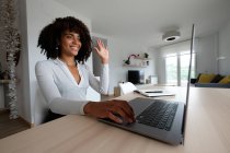 Alegre freelancer afroamericana hablando en video chat y saludando con la mano mientras trabaja desde casa y usando netbook - foto de stock