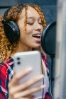 Alegre cantante afroamericana femenina en auriculares con smartphone interpretando canción contra escudo de sonido en estudio de grabación - foto de stock
