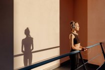 Mujer joven apoyada en la barandilla y escuchando música usando el teléfono después de hacer ejercicio al aire libre al atardecer - foto de stock
