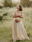 Спокойная беременная женщина, покрывающая голые груди, стоит в сельской местности летом и смотрит в сторону — стоковое фото
