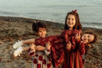 Lindas niñas étnicas sosteniendo hermanita en las manos mientras se divierten juntos en la playa de arena cerca del mar - foto de stock