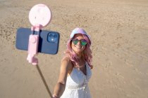 Hochaufragende Frau mit rosa Haaren und sommerlichem Outfit, die an einem sonnigen Tag am Strand am Selfie-Stick ein Selfie mit dem Smartphone macht — Stockfoto