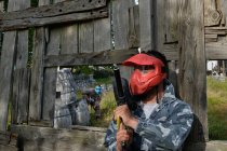 Männlicher Spieler in Maske und mit Paintballpistole, der während des Spiels hinter einem Holzzaun steht — Stockfoto
