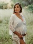 Транквіль вагітна жінка в одязі, зворушлива животик з закритими очима, стоячи в полі в сільській місцевості в літній день — стокове фото
