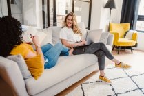 Frohes lesbisches Paar blättert im Netbook und liest interessante Bücher, während es sich auf der bequemen Couch im modernen Wohnzimmer ausruht — Stockfoto