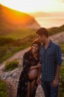 Feliz mulher grávida hispânica e homem amoroso de mãos dadas e abraçando uns aos outros enquanto caminham ao longo da costa montanhosa na noite de verão — Fotografia de Stock
