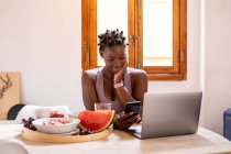 Encantada afro-americana navegando telefone celular enquanto se senta à mesa com frutas maduras saudáveis em casa — Fotografia de Stock