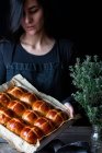 Жінка-пекар, що тримає пух, свіжоспечений гарячий хрест булочки на випічці лотка — стокове фото