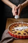 Anonymous person in apron standing at wooden table and sprinkling sugar powder on top of delicious homemade pie with cherries — Stock Photo