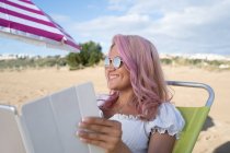 Joyeuse indépendante assise dans une chaise longue et travaillant sur une tablette sur une plage de sable près de la mer en été — Photo de stock