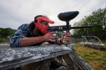 Male player in camouflage outfit and with gun lying down on abandoned car and aiming while playing paintball in countryside — Stock Photo